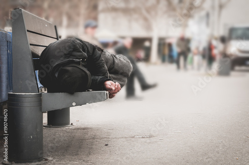 Poor homeless man or refugee sleeping on the wooden bench on the urban street in the city, social documentary concept, selective focus photo