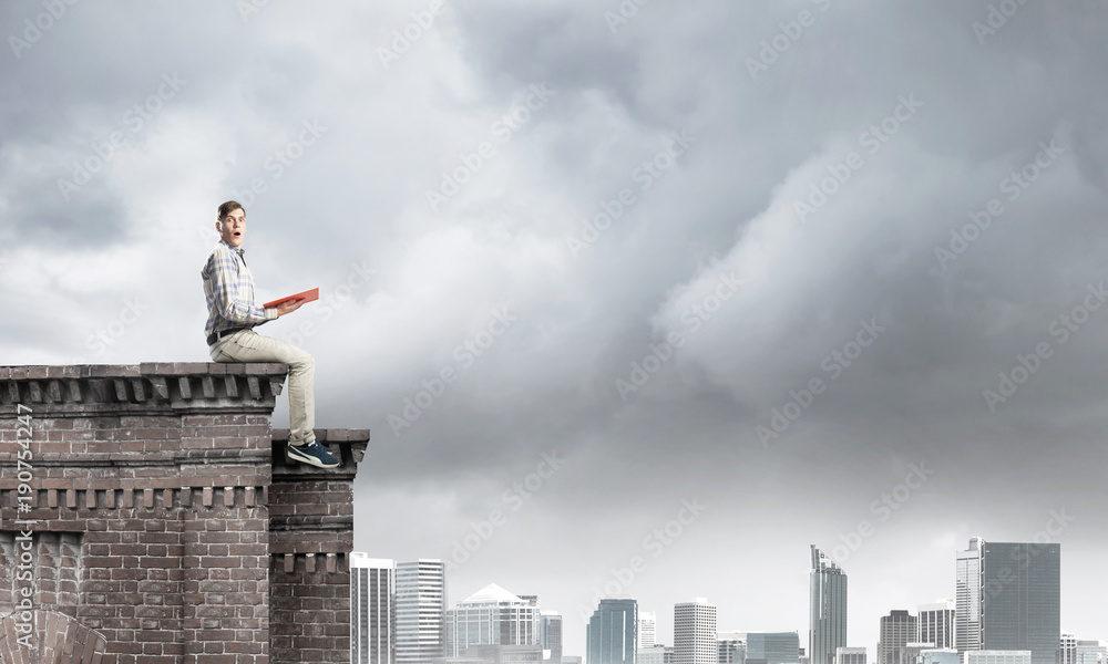 Man on roof edge reading book and cityscape at background