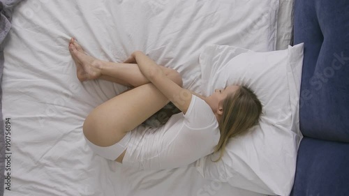 Sad depressed young woman with blond hair lying in fetal position on bed and crying. Lonely unhappy attractive female holding her knees with arms, sobbing and crying alone in bed