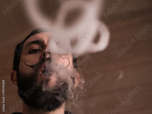 Man with Beard and Mustages Vaping an Electronic Cigarette. Vaper Hipster Smoke Vaporizer and Exhals Smoke Rings.