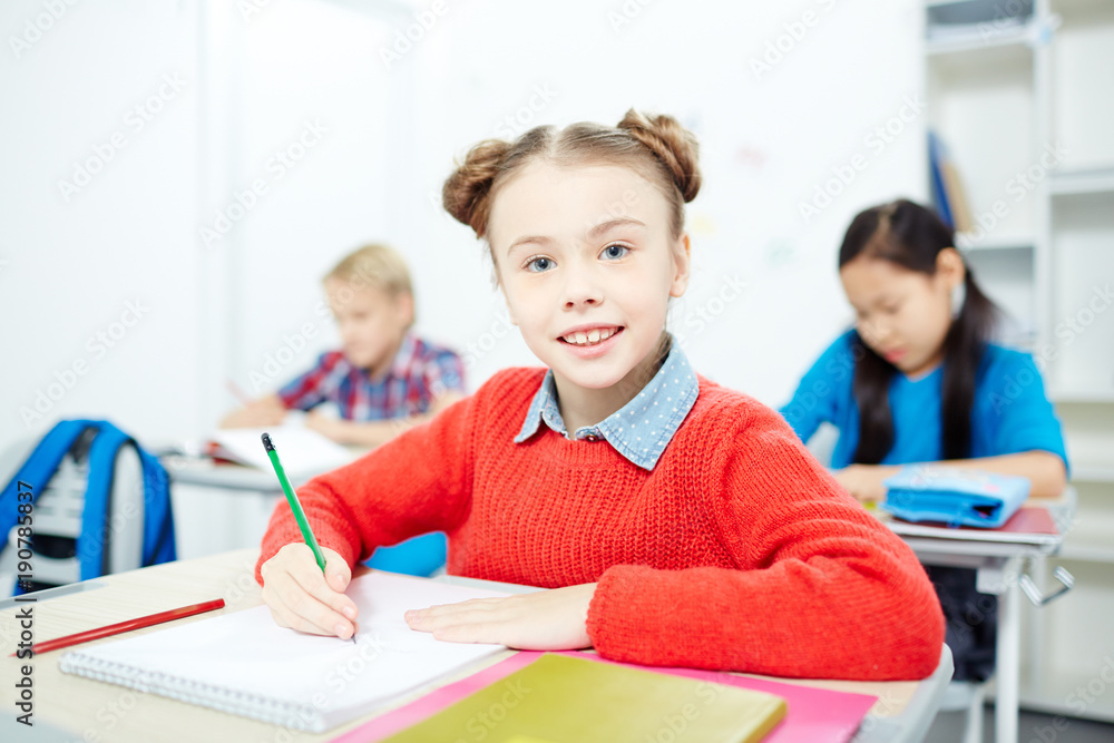 Learner of elementary school sitting by desk, drawing with crayons and looking at camera Learner of elementary school sitting by desk, drawing with crayons and looking at camera