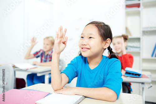 Adorable schoolgirl and her classmates raising their hands while working at lesson