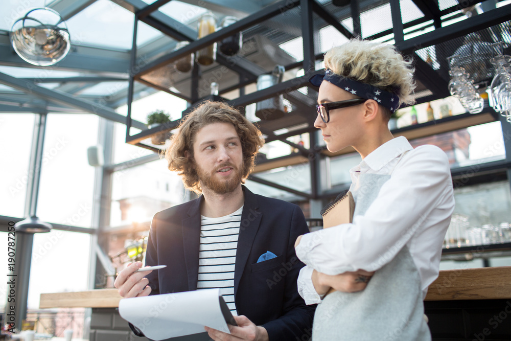 Young confident revision manager or owner of cafe having talk to new waitress at the beginning of her first working day Young confident revision manager or owner of cafe having talk to new waitress at the beginning of her first working day