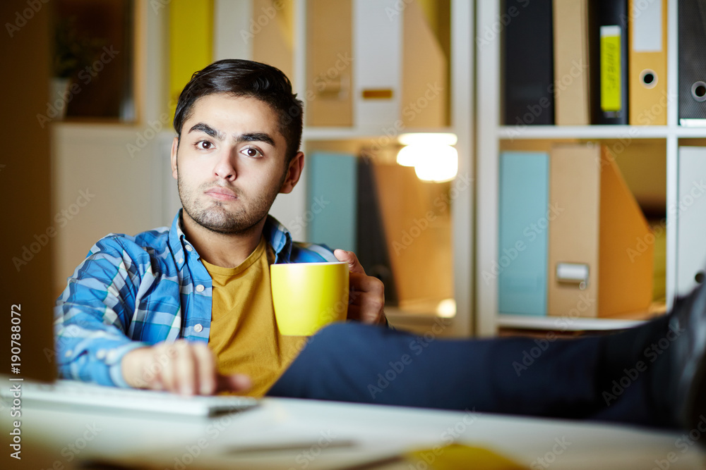 Pensive young man with cup of tea or coffee looking at camera while relaxing in front of desktop monitor Pensive young man with cup of tea or coffee looking at camera while relaxing in front of desktop monitor
