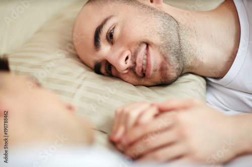 Young smiling man with his head on pillow looking at his partner in bed