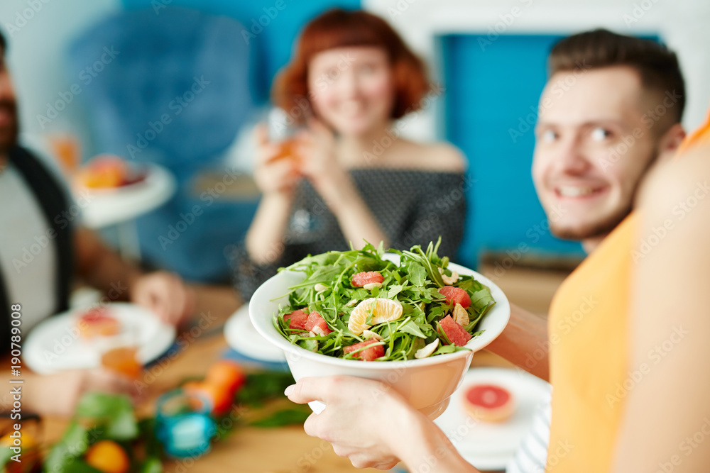 Woman holding bowl of fresh vegetarian salad with greenery, tomatoes, mandarines and ruccola while her guests waiting for festive dinner