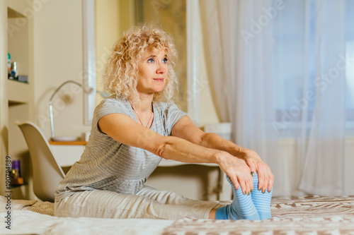 Lazy gymnastics on the bed concept - middle aged curly blond caucasian woman in pajamas sitting on the bed and stretching her body after sleep in early morning. photo