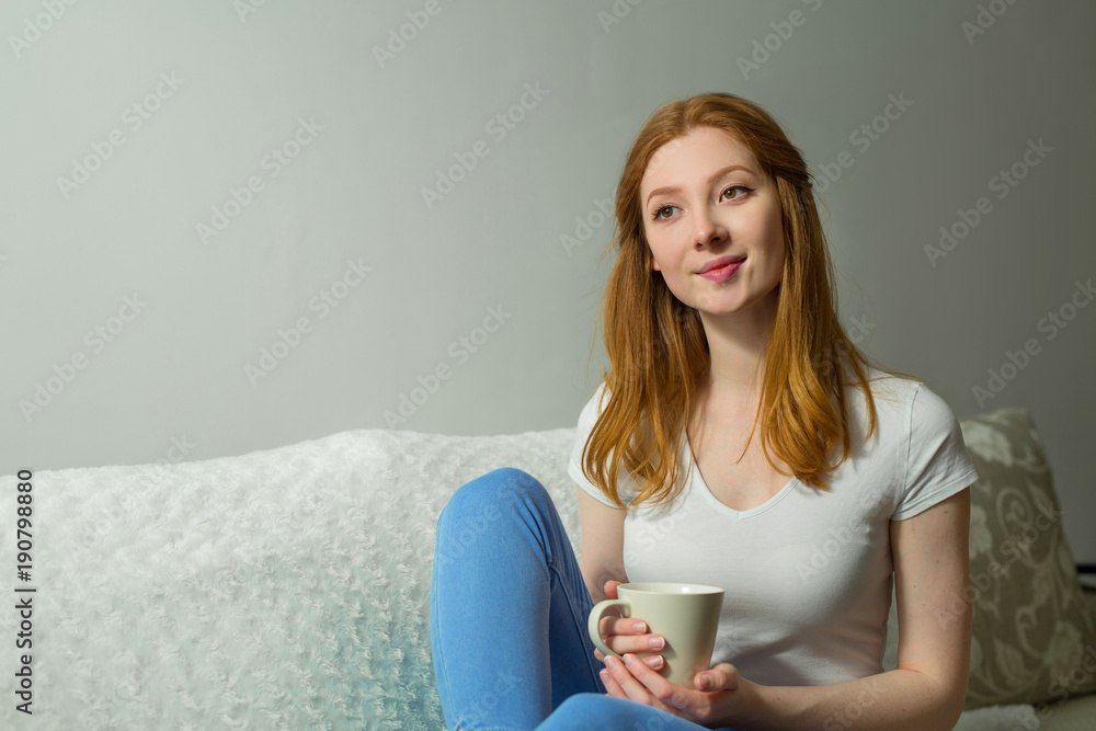 beautiful young girl with red hair sitting on the couch with a cup of tea beautiful young girl with red hair sitting on the couch with a cup of tea