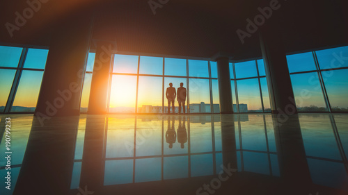 The two men stand near the panoramic window on the sunset background