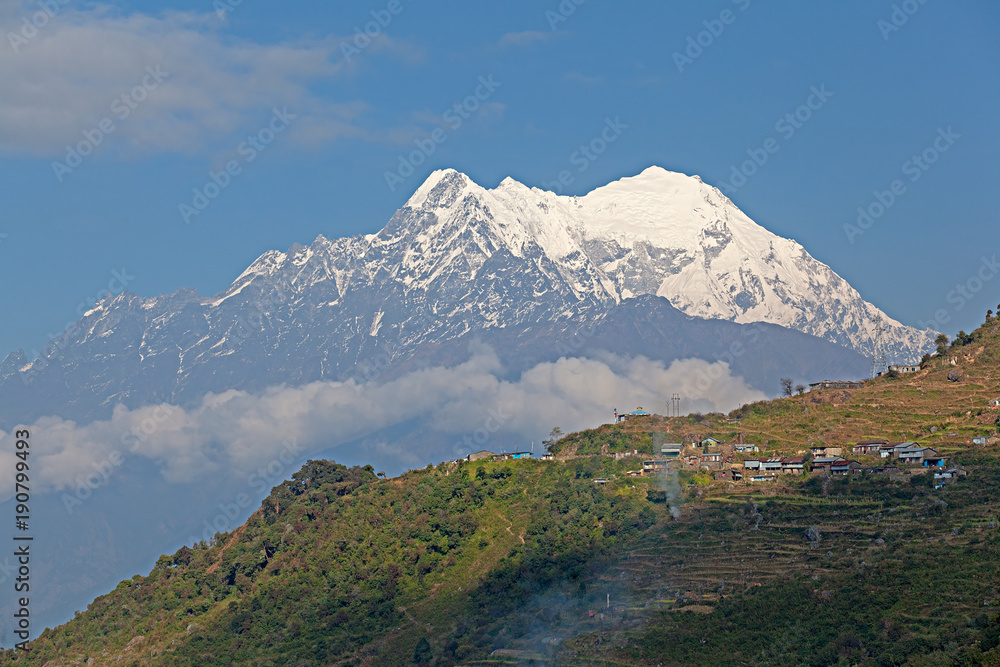 Himalayan mountains with green wooded mountain slope with a small village on it in the foreground and a big showcapped peak of Manasly Himal mountain massif in clouds in the background, Lantang, Nepal