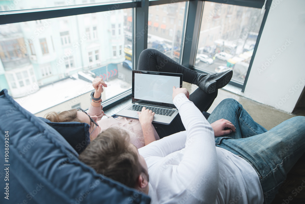 Young man with a girl working on a laptop at the window on the couch. Workers lie on the couch and work. Young man with a girl working on a laptop at the window on the couch. Workers lie on the couch and work.