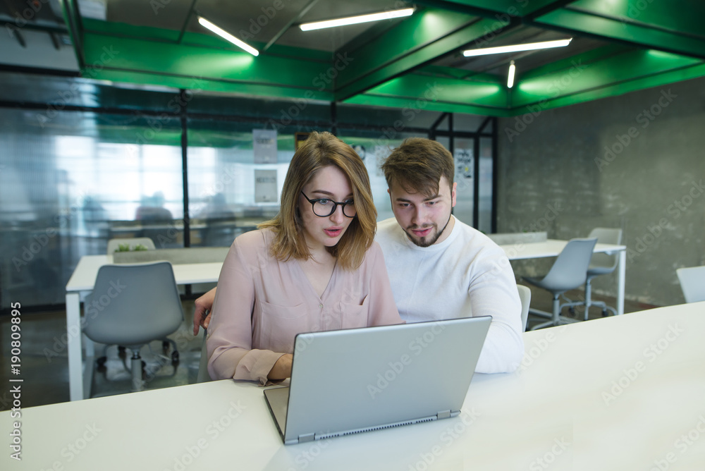 Beautiful young couple working with a laptop in a stylish, modern office. A man and a girl are looking at the notebook monitor in the office. Beautiful young couple working with a laptop in a stylish, modern office. A man and a girl are looking at the notebook monitor in the office.