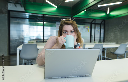 Beautiful girl sitting at the desk in the office and drinking coffee from the cup while working on a computer. Office work concept.