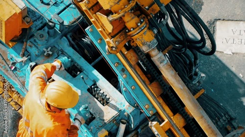 Man dressed in yellow helmet and working clothes operating metal drilling rig using the functional buttons. Worker controlling the hard rotary pipe before making the well