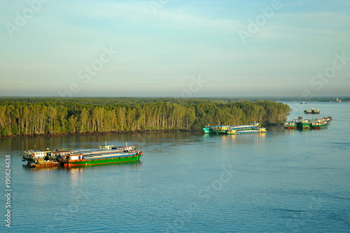 Cargo boats on a river near Ho Chi Minh City. A forest runs down to the water's edge. The sky is blue, with shite clouds. The water is luminescent.