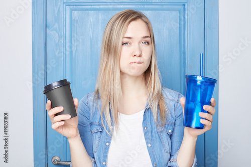 .Doubtful hesitant intriguing blond female curves lips, has thoughtful expression, can`t decide or make choice between addiction and health, dressed in blue casual shirt. Facial expressions concept.