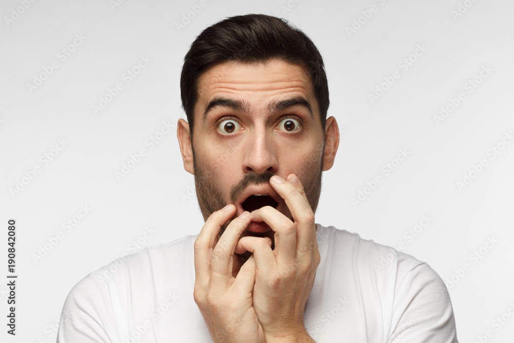 Young man isolated on grey background dressed in casual clothes, covering mouth with hands and round eyes experiencing deep astonishment and fear Young man isolated on grey background dressed in casual clothes, covering mouth with hands and round eyes experiencing deep astonishment and fear