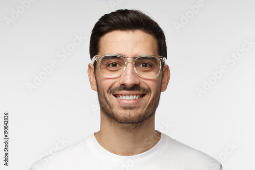 Close up shot of smiling attractive man in white t-shirt and trendy trasparent eyeglasses isolated on gray background © Damir Khabirov