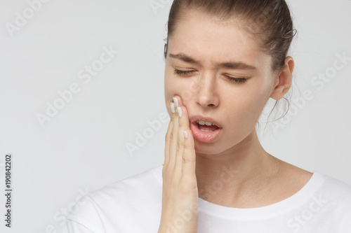Young European woman isolated on grey background suffering from severe toothache  feeling pain so strong that she is pressing fingers to cheek to calm it down  looking desperate