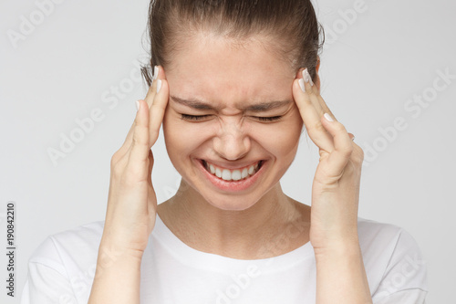 Portrait of good looking young woman isolated on grey background suffering from severe headache, pressing fingers to temples, closing eyes in order to relieve pain with helpless face expression