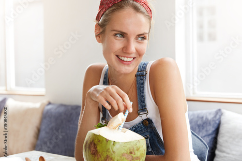 Pleased cheerful young female with positive smile sips exotic coconut cocktail, rests at comfortable couch, looks aside, thinks about something pleasant, dreams happily, glad have vacations. photo