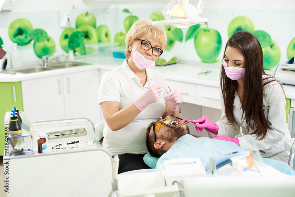 Fototapeta premium Attractive female dentist is smiling to the camera while holding medical instruments getting ready to attending the teeth of a male client of a dental office. Young brunette is assisting the doctor.