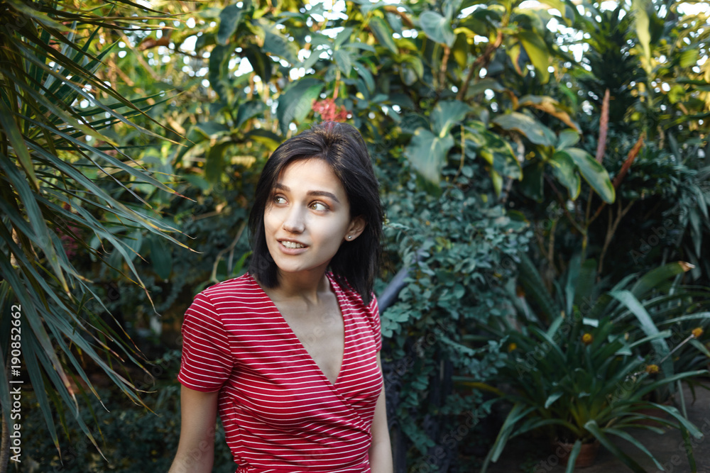 Picture of attractive positive young Caucasian woman with brunette hair walking in tropical forest, hiding herself in shadow of exotic trees on hot sunny day, looking sideways with curious expression Picture of attractive positive young Caucasian woman with brunette hair walking in tropical forest, hiding herself in shadow of exotic trees on hot sunny day, looking sideways with curious expression