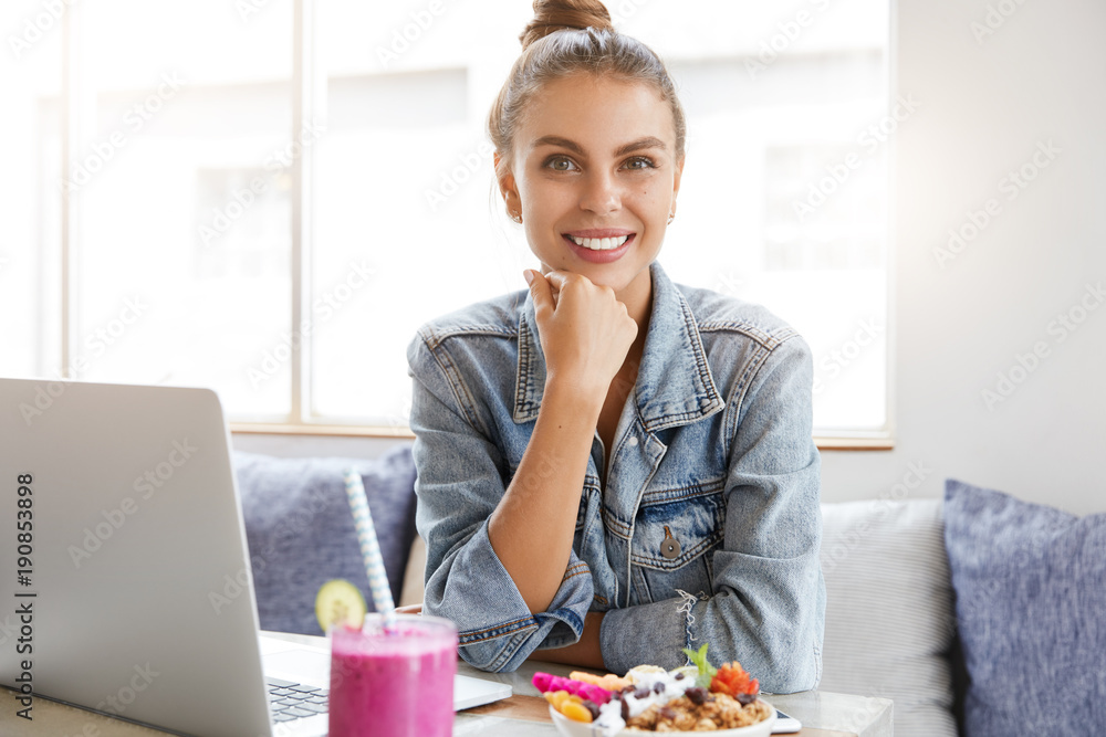 Satisfied cheerful female with hair bun, dressed in denim jacket, enjoys weekend, watches favourite film on laptop computer, tastes delicious fruit salad and drinks fresh smoothie, relaxes at couch
