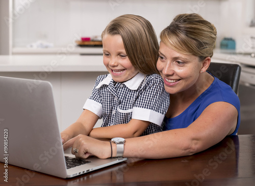 blond mother woman together with her young beautiful and sweet little girl 6 to 8 years old sitting at home kitchen enjoying with laptop computer