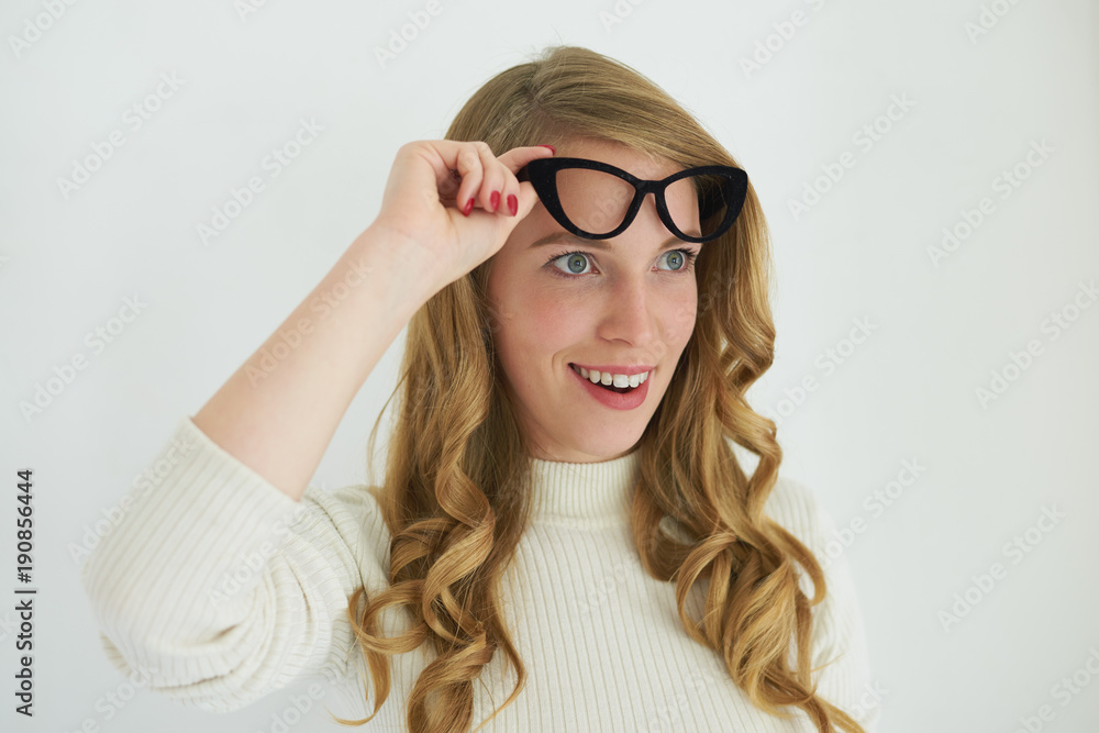 Horizontal shot of attractive young female wearing white turtleneck smiling excitedly, raising her stylish cat eye glasses and looking ahead of her in astonishment, shocked with some unexpected news