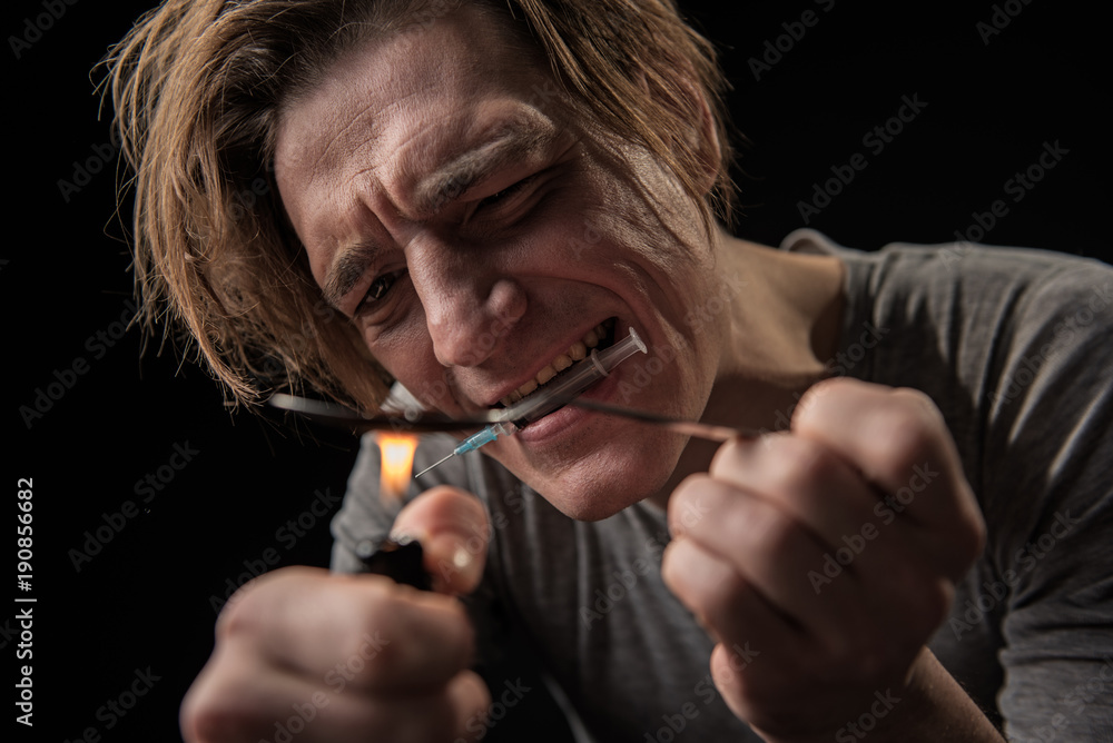 Fototapeta premium Young sad narcomaniac preparing dose of hop. He is holding syringe with teeth. Focus on male expression. Isolated on background