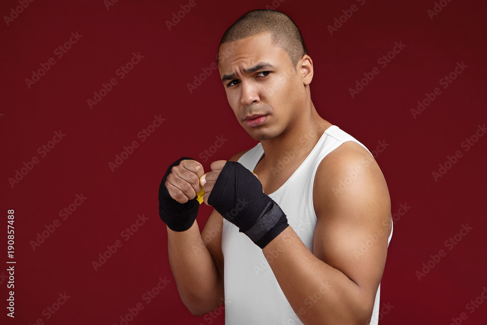 Isolated studio shot of furious young Afrio American male fighter wearing white sleeveless sports shirt and black boxing bandages, looking at camera with fierce look while working out in gym
