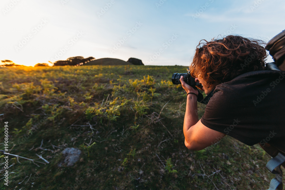 Young photographer with long brown curly hair, tattoo on his wrist and camera backpack on his back is taking picture of a sunset during golden hour in the mountains of Madeira with grove in background