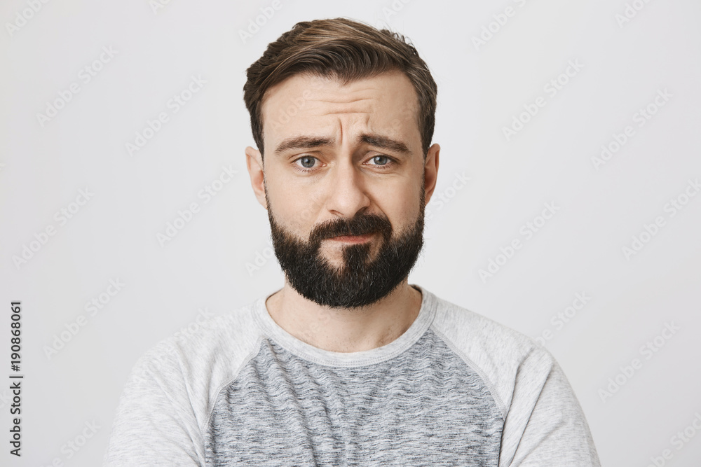 Studio portrait of uncertain and disappointed bearded male adult, looking at camera with sad look and frowned eyebrows, standing against gray background. Man feels stomach ache during shift Studio portrait of uncertain and disappointed bearded male adult, looking at camera with sad look and frowned eyebrows, standing against gray background. Man feels stomach ache during shift