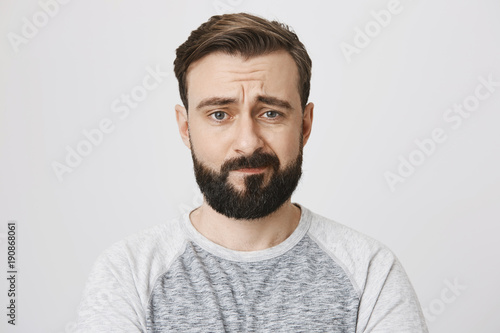 Studio portrait of uncertain and disappointed bearded male adult, looking at camera with sad look and frowned eyebrows, standing against gray background. Man feels stomach ache during shift
