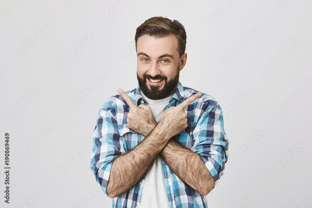 Indoor portrait of excited young adult with beard and trendy haircut, pointing in different directions with crossed arms, over gray background. Man with tricky smile trying to puzzle his friend Indoor portrait of excited young adult with beard and trendy haircut, pointing in different directions with crossed arms, over gray background. Man with tricky smile trying to puzzle his friend