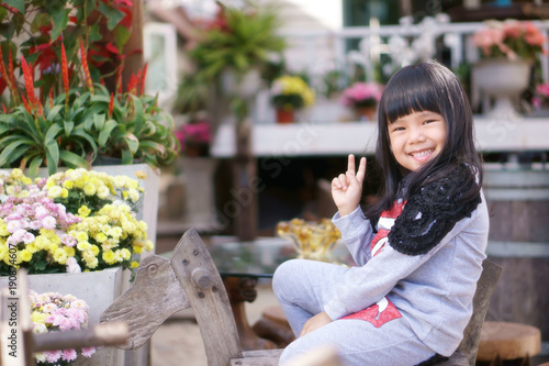 Asian children cute or kid girl happy fun with smile and show two finger in beautiful flower garden and sit on wooden horse chair for relax on holiday travel at vintage white house or cafe restaurant © kornnphoto