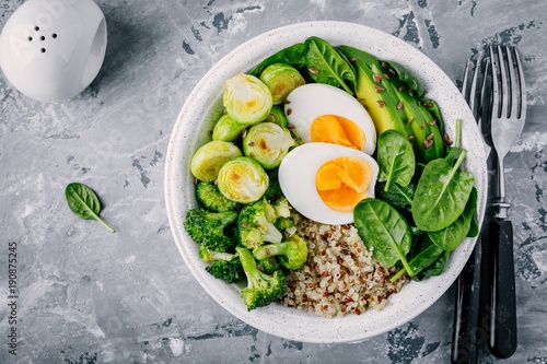 Healthy green vegetarian buddha bowl lunch with eggs, quinoa, spinach, avocado, grilled brussels sprouts and broccoli on dark grey background.