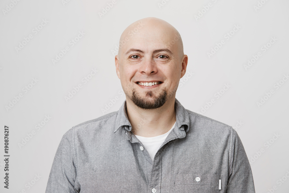 Close-up portrait of positive and attractive bald caucasian adult man smiling cheerfully while standing over gray background. Guy quit his job to start traveling across world so he feels happy Close-up portrait of positive and attractive bald caucasian adult man smiling cheerfully while standing over gray background. Guy quit his job to start traveling across world so he feels happy
