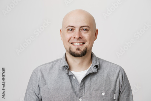 Close-up portrait of positive and attractive bald caucasian adult man smiling cheerfully while standing over gray background. Guy quit his job to start traveling across world so he feels happy
