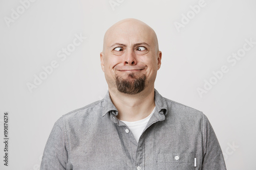 Portrait of funny and childish bald caucasian male model making crazy eyes and smiling strangely, standing over gray background. Guy shows daughter that he can see his own nose without mirror