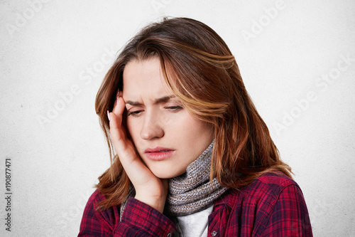 Illness and sickness concept. Exhausted stressed young female suffers from migraine, keeps hand on head, looks desperately down, isolated over white background. Woman has terrible headache