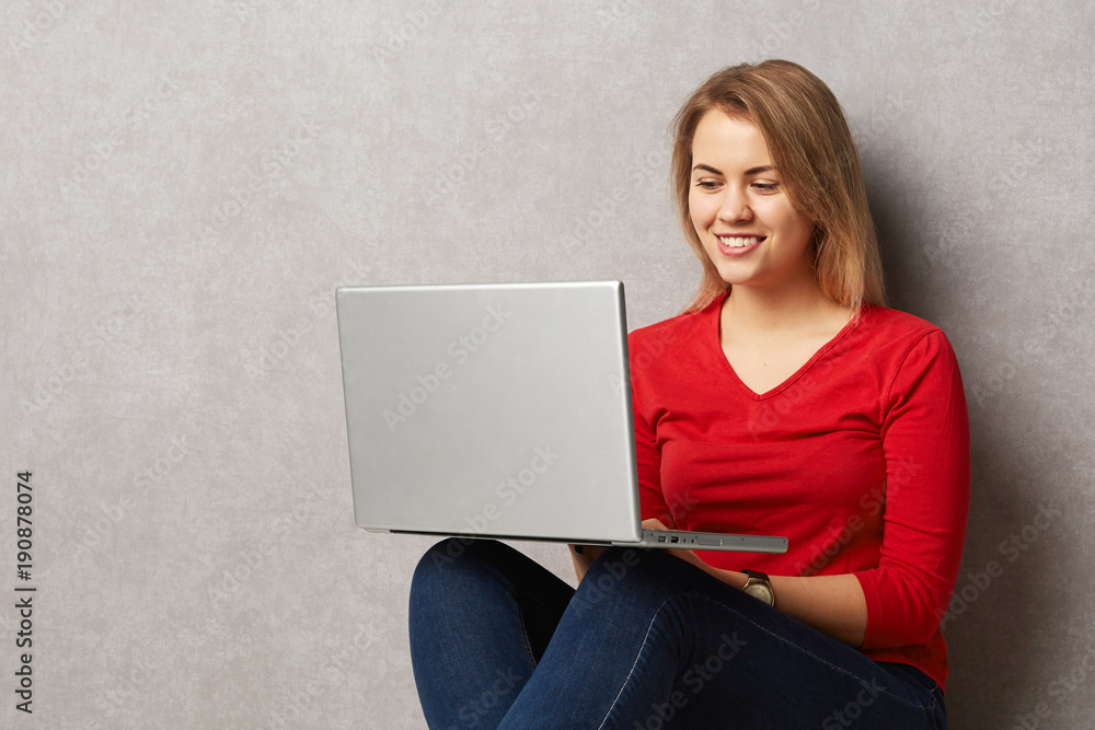 Beautiful pleased young female sits on floor with digital laptop computer, enjoys online communication, being always in touch, isolated over grey background. Woman surfes webpages in internet Beautiful pleased young female sits on floor with digital laptop computer, enjoys online communication, being always in touch, isolated over grey background. Woman surfes webpages in internet
