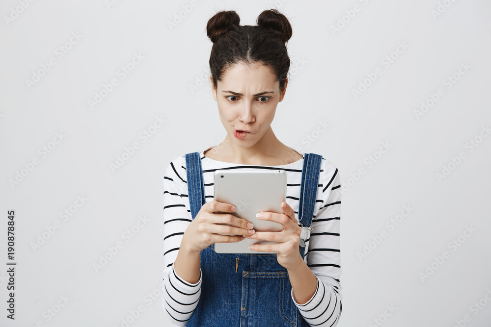 Portrait of cute european girl with trendy two buns hairstyle, wearing overalls and looking excited or interested at tablet screen, standing over gray background. Woman looks who liked her boyfriend Portrait of cute european girl with trendy two buns hairstyle, wearing overalls and looking excited or interested at tablet screen, standing over gray background. Woman looks who liked her boyfriend