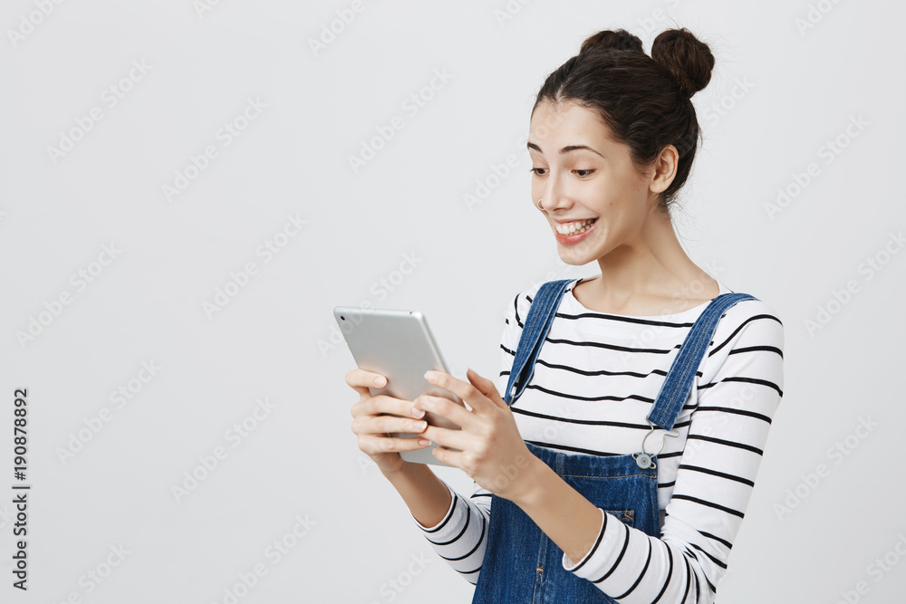 Fototapeta premium Half-turned european model with two buns hairstyle and pierced nose smiling cheerfully while holding tablet and browsing through it, standing in overalls against gray background.
