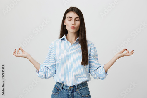 Young and attractive successful female standing with closed eyes and folded lips while spreading hands and showing zen sign as if doing yoga, over gray background. Sportswoman tries stay calm