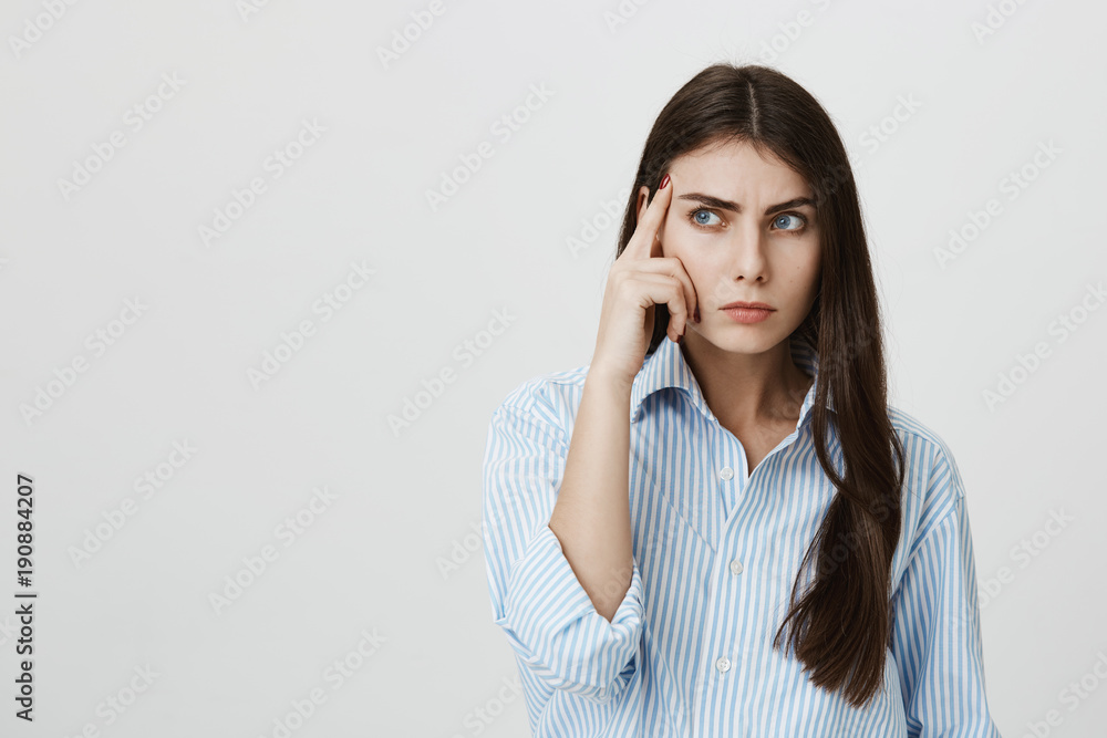 Young attractive european female adult with serious expression, thinking about something or calculating in mind while holding hand on temple, standing over gray background. Student can not recall info Young attractive european female adult with serious expression, thinking about something or calculating in mind while holding hand on temple, standing over gray background. Student can not recall info