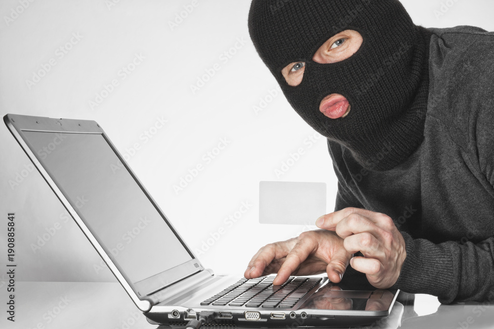 Hacker in balaclava holding a card in left hand and typing something with right hand on laptop keyboard Hacker in balaclava holding a card in left hand and typing something with right hand on laptop keyboard