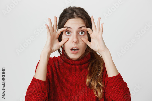 Dark-haired female dressed in casual clothes with opened mouth and popped eyes looks at camera with stunning expression of face. Amazed shocked woman with bugged eyes poses indoors photo