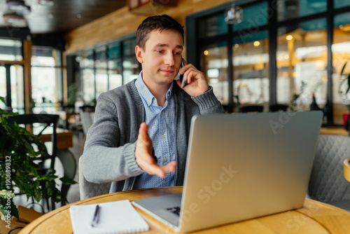 Young handsome man sitting in co-working office, talking on phone, casual style, confident, working on laptop online, freelancer, busy